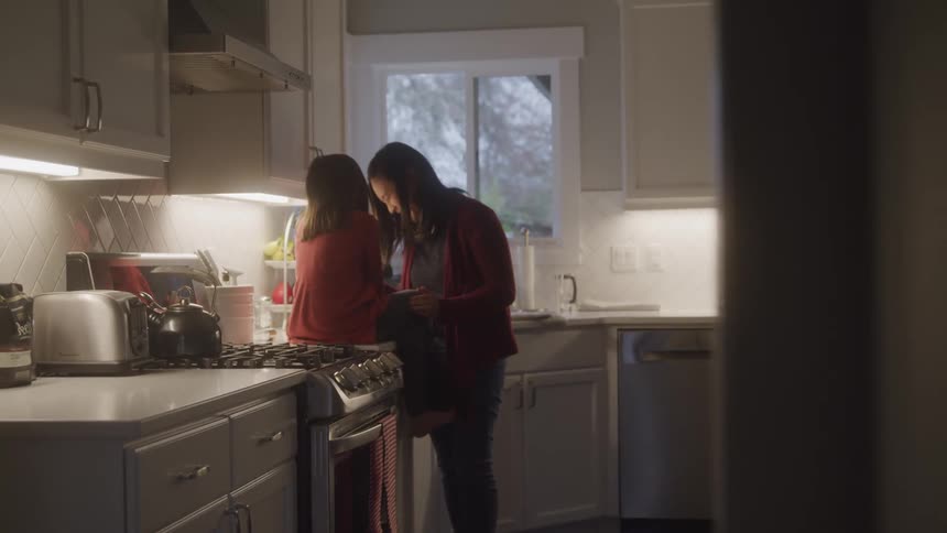 A woman standing on a kitchen smiling at a little girl who is sitting on the counter next to the stove