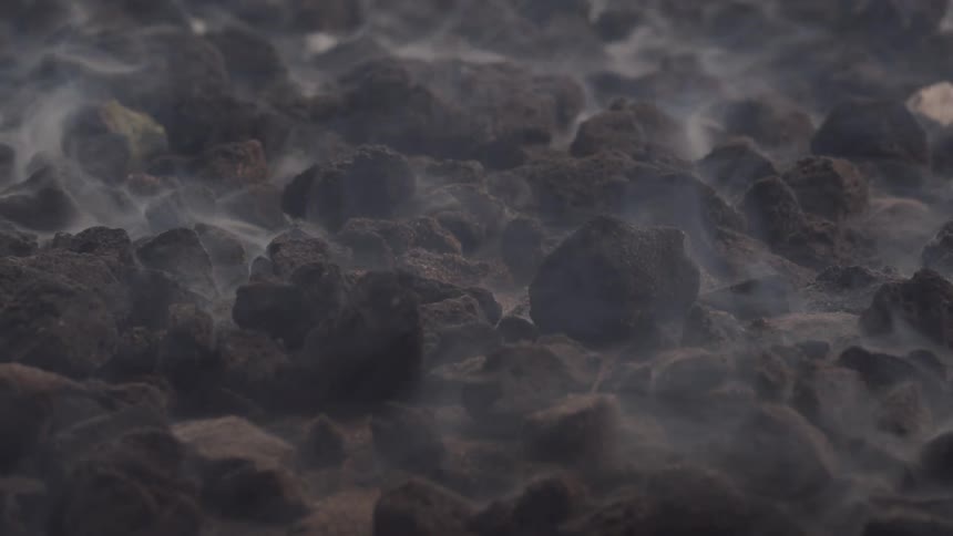 A rugged dark rocky surface with light mist or steam gently rising suggesting geothermal activity or heat