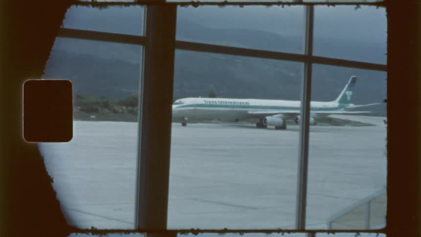 A vintagestyle shot shows a jet airliner parked on an airport tarmac viewed through the large windows of an airport terminal