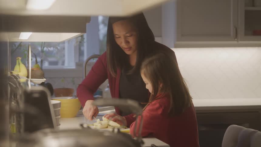 A child assist an adult in cutting vegetables on a wooden countertop in a bright kitchen with white cabinets and a tiled backsplash