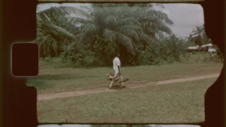 A man walks along a dirt path carrying a bundle of sticks in a lush green outdoor setting captured on vintage film