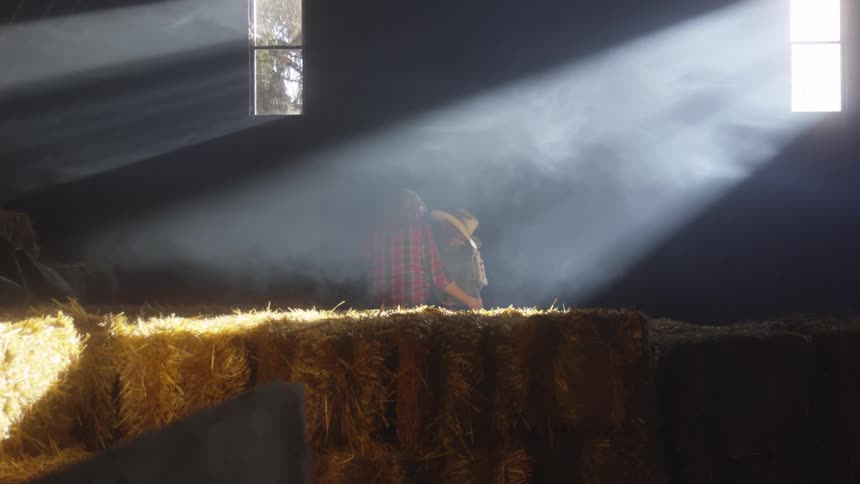 A couple sits on hay bales inside a barn illuminated by sunlight streaming through a window