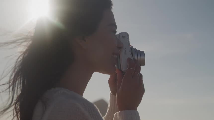 A young woman with flowing hair stands against a bright sky holding a camera to her face capturing a moment with a serene and artistic expression