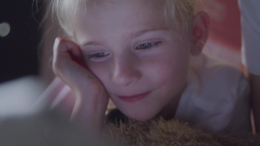 Portrait of a little blonde girl lying down with a happy facial expression and interacting with a screen