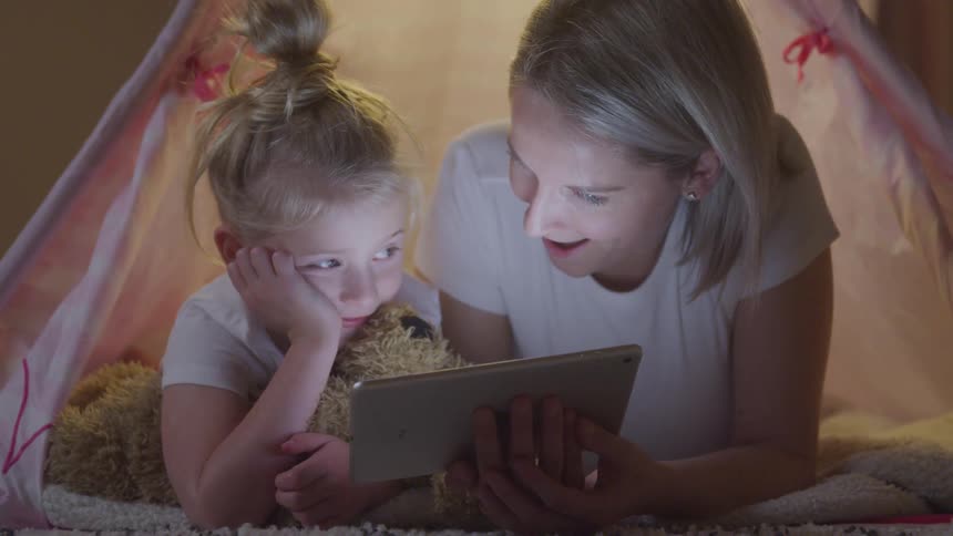 Mother and daughter lie in a tent setup indoors and share a tablet