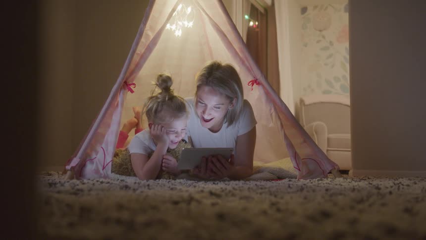 Mother and daughter lying together in a tent on the living room carpet and interacting with a tablet happily