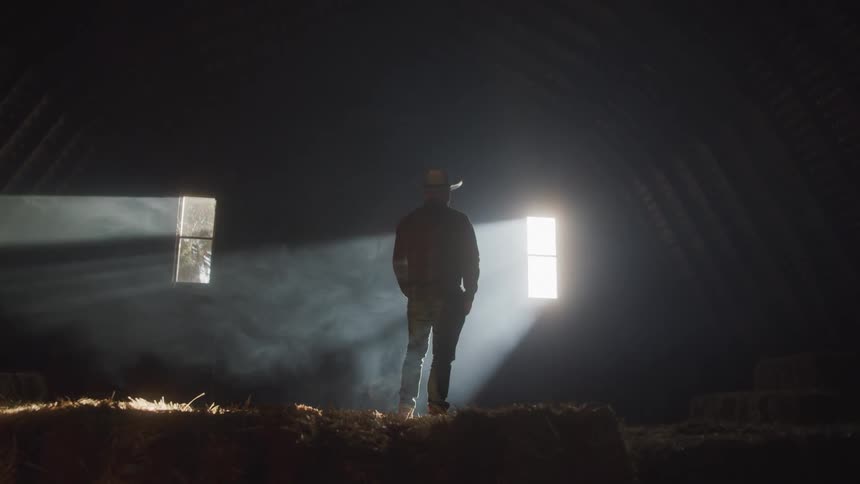A lone figure in a cowboy hat stands in a dark barn illuminated by shafts of light streaming through windows
