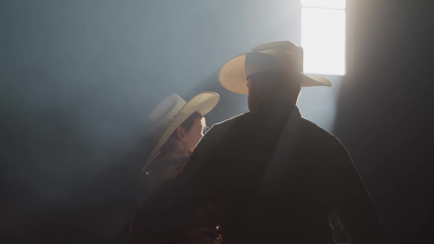 A father and son sit next to each other inside of a dark barn