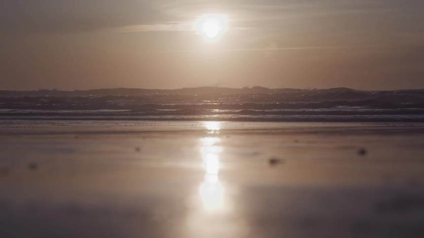 A low angle shot shows the sun reflecting on the wet sand of a beach with gentle waves during a sunset