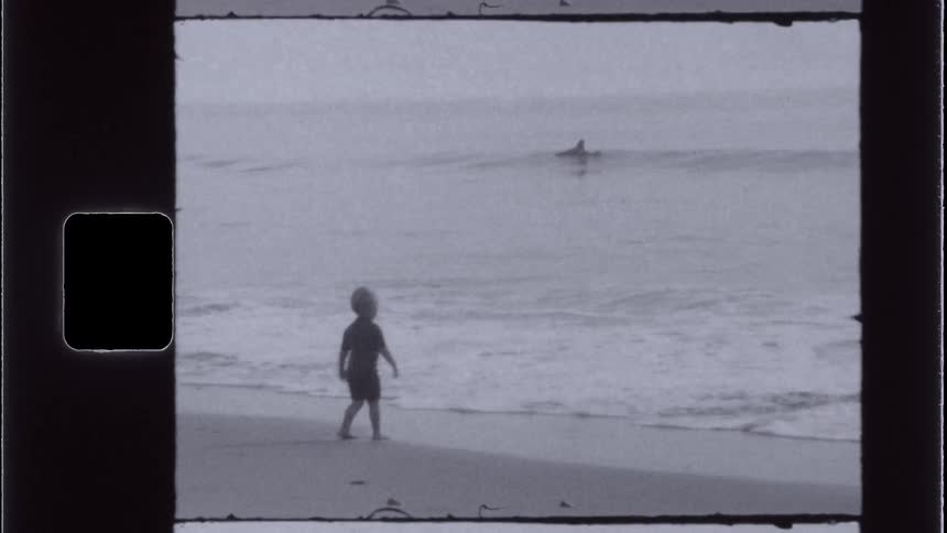 A grainy black and white film shot captures a young child standing at the shoreline gazing at something in the water