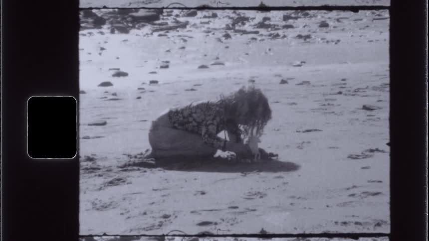 A vintagestyle shot shows a person kneeling on a sandy surface possibly a beach with their head down and long hair obscuring their face