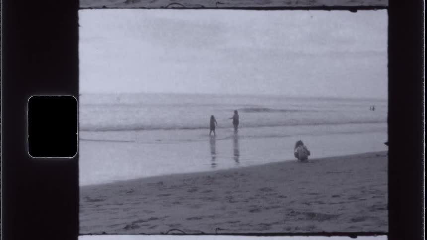 A girl plays in the sand on a beach while a woman and another girl walk through the shallow water nearby