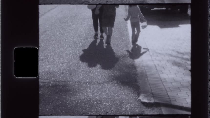 A vintagestyle shot shows people walking away from the camera on a paved surface with their shadows visible in the foreground