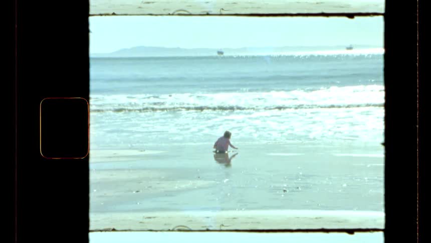 A girl sits at the edge of a beach with waves rolling in and a distant shoreline visible