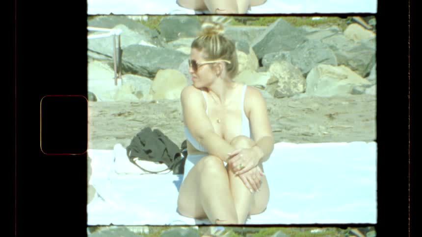 A blonde woman wears a bikini while sitting on a white beach blanket on a summer day