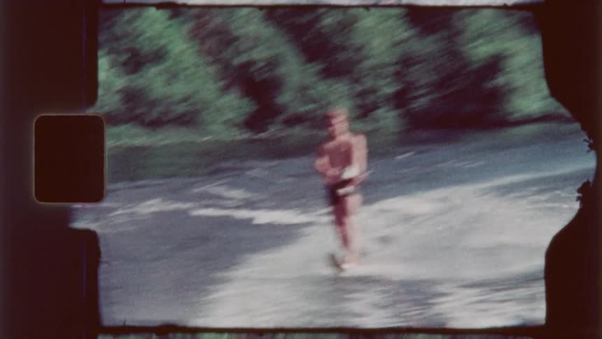 Man water skis behind a boat passing through the waters near a forested shore during the daytime