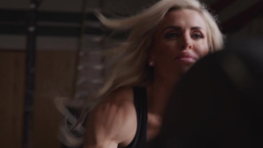 A woman with blonde hair uses a rowing machine in a gym with visible effort shown in her arm muscles