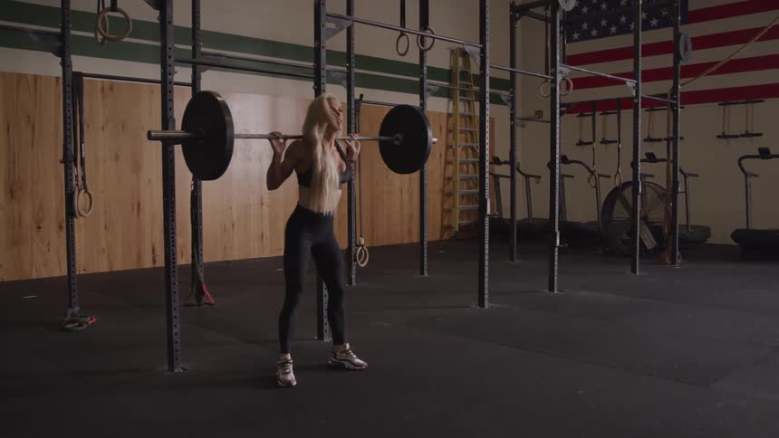 A fit woman with long blonde hair performs a squat with a barbell in a gym setting