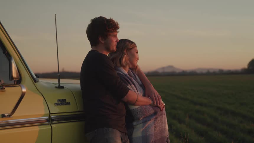 A man and woman embrace as they lean on a truck and look into the distance