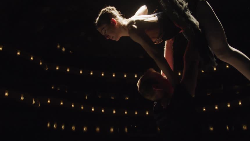 A ballet dancer in a black tutu is lifted gracefully by a partner on a dimly lit stage with rows of lights forming a glowing backdrop