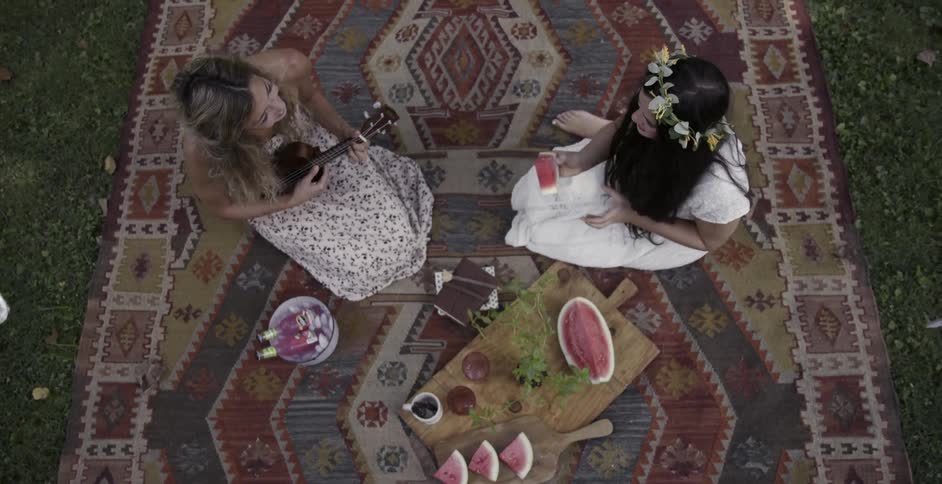 Two women in dresses sit on a carpet in the grass
