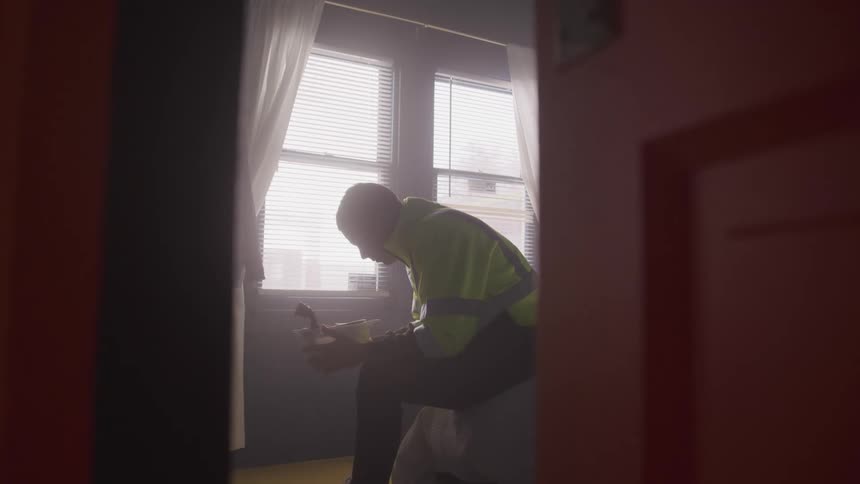 A man wearing a green jacket is sitting on a bed in a bedroom holding a toy