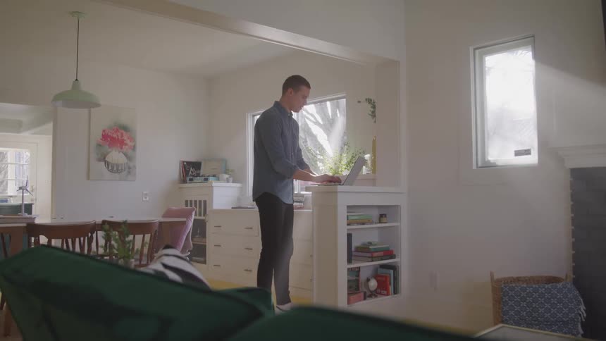 A person stands at a kitchen counter using a laptop with a dining area in the background and a bookshelf on the side