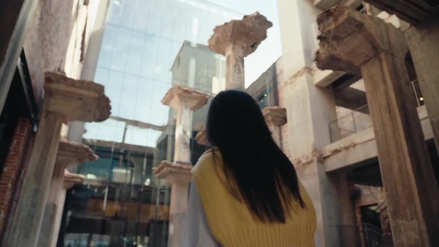 A woman in a lightcolored outfit walks around in a courtyard surrounded by weathered columns and modern buildings looking upwards