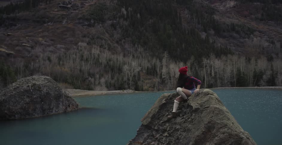 A view of a large mountain with some water in front and some rocks with a girl sitting on the rocks