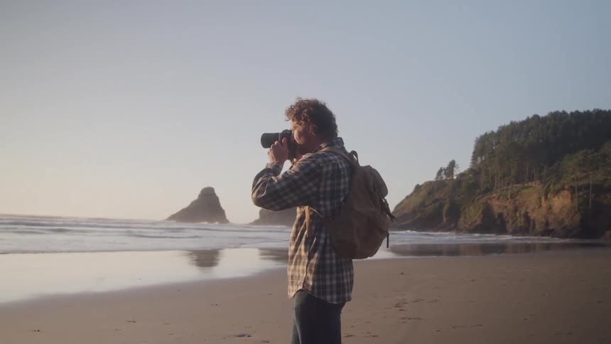 A person in a plaid shirt with a backpack photographs the ocean and rock formations from a sandy beach with forested cliffs in the background