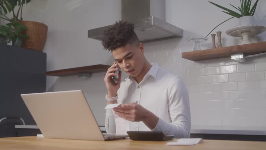 A young man reads a note while sitting at a desk and talking on his smartphone