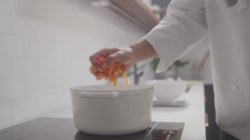 A chef in their white uniform in a kitchen takes a handful of bright orange carrots from a bowl and adds them to a white pot already placed on the electric stovetop