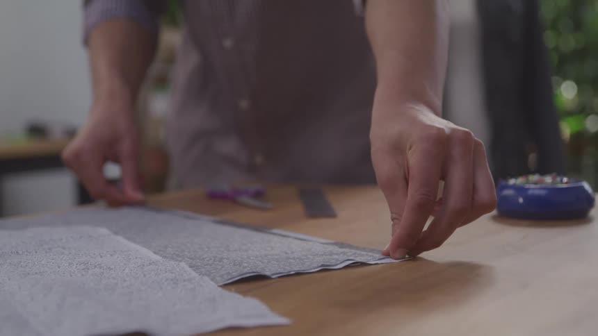 Hands measure and manipulate a piece of lightcolored fabric on a wooden table with tools nearby