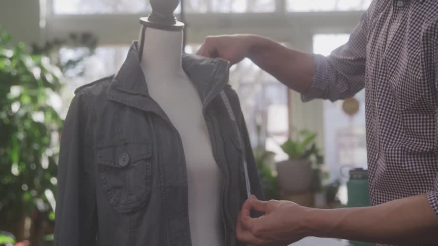 In a workshop setting a persons hands are visible using a measuring tape to take measurements of a jacket on a mannequin with various plants situated in the area near the windows in the background