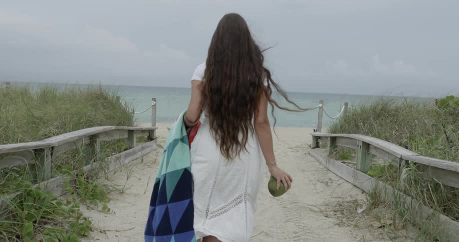 Woman walking on sandy beach path holding towel and coconut