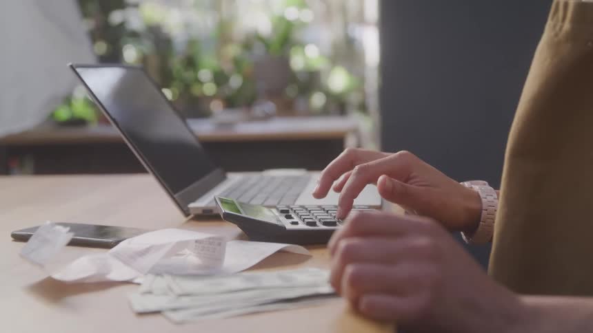 Hand typing on a calculator on a table next to a laptop and receipts