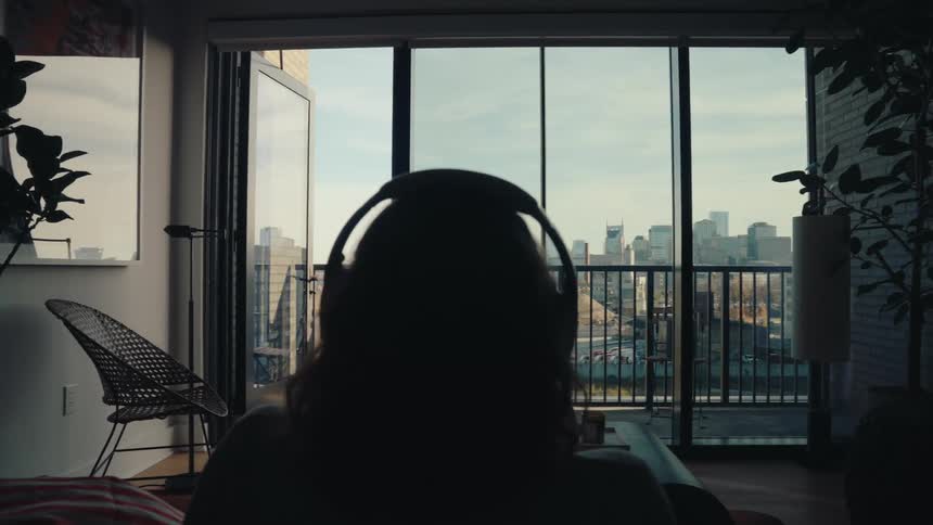 A person wearing headphones sits facing a balcony with a city view seen from inside an apartment