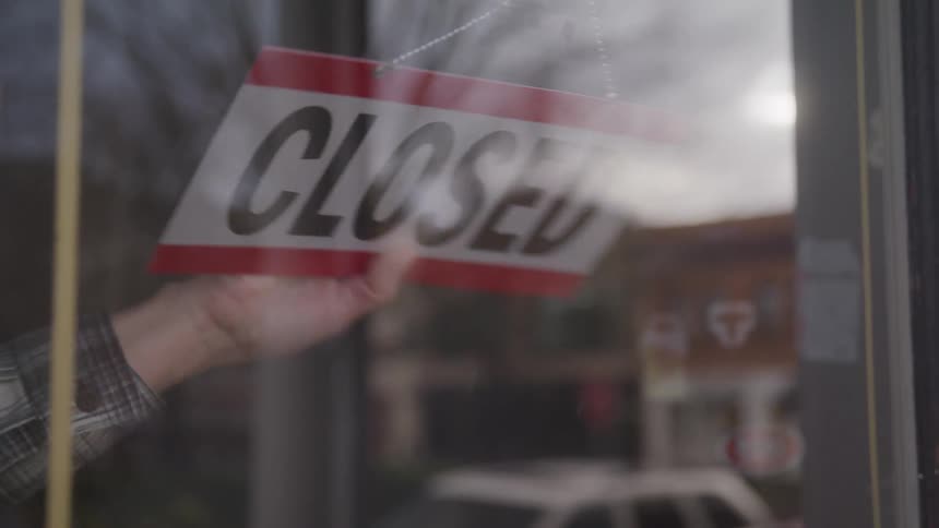 A hand flips an open sign in a storefront window with a street scene faintly reflected in the glass
