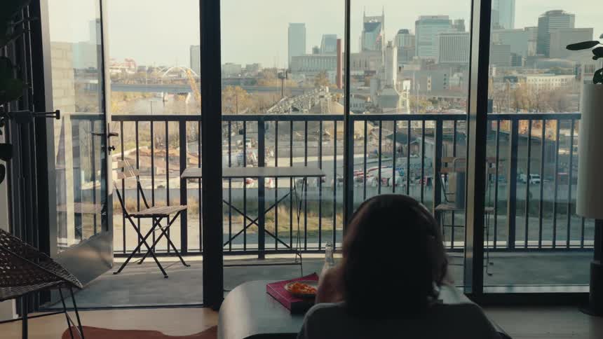 Woman reads a book while sitting in her living room with a view of the city skyline and the nearby river on an autumn day