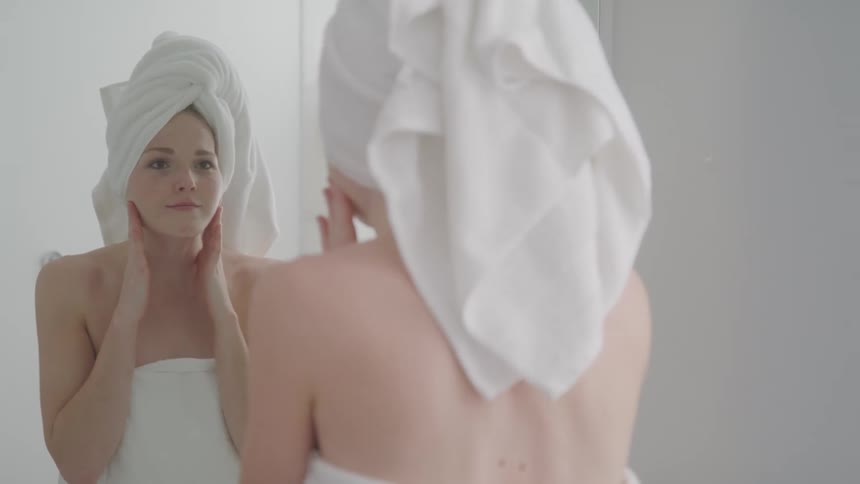 A woman with a towel wrapped around her head and body looks at her reflection in a mirror touching her face