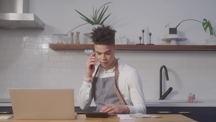 A young man sits at a desk while talking on his smartphone and using a calculator