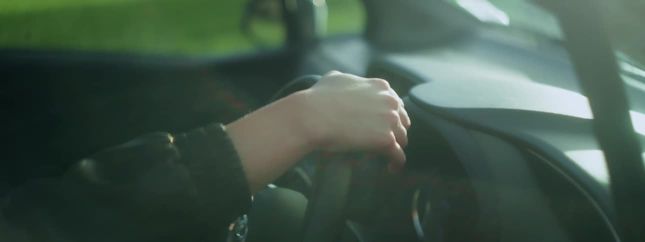 A persons hand grips a cars steering wheel from the inside at morning