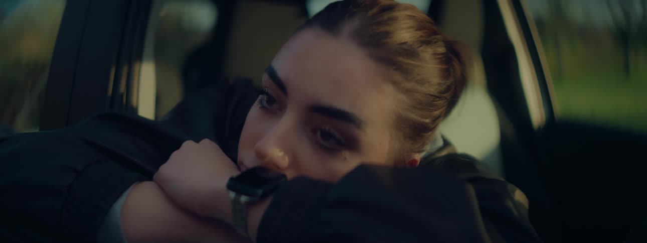 A woman with blonde hair is sitting in a parked car resting her head on her arms looking out the window