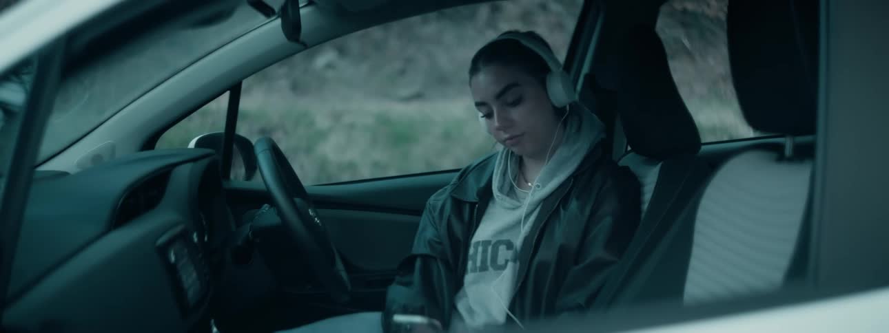 A young woman with headphones rests in a car gazing downward at her phone