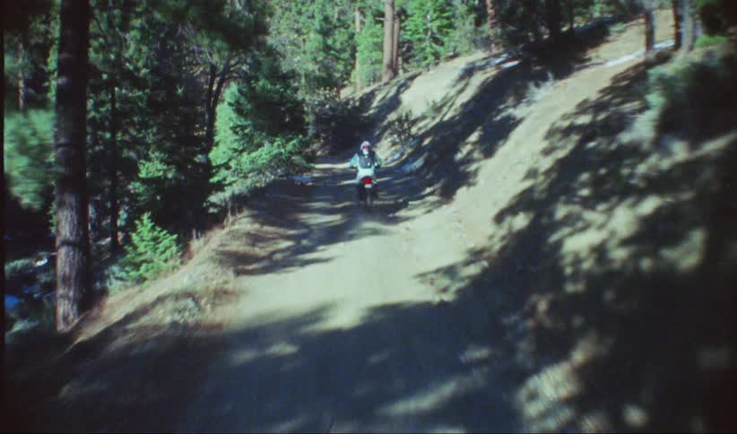 A person rides a dirt bike on a remote road in a forested mountain range on a clear day