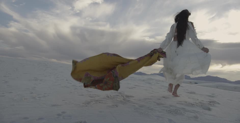 A woman holding a long scarf walks across a sandy landscape on a windy day