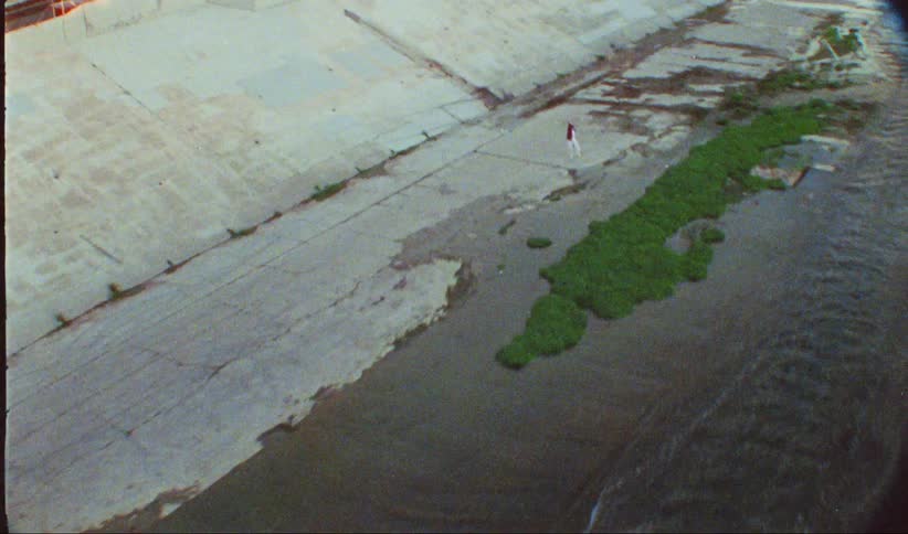 A woman in white pants and a red jacket dances on cracked concrete within a shallow drainage canal