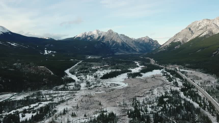 Aerial view of a snow covered valley surrounded by towering mountains with a winding river and a road cutting through the scenic landscape