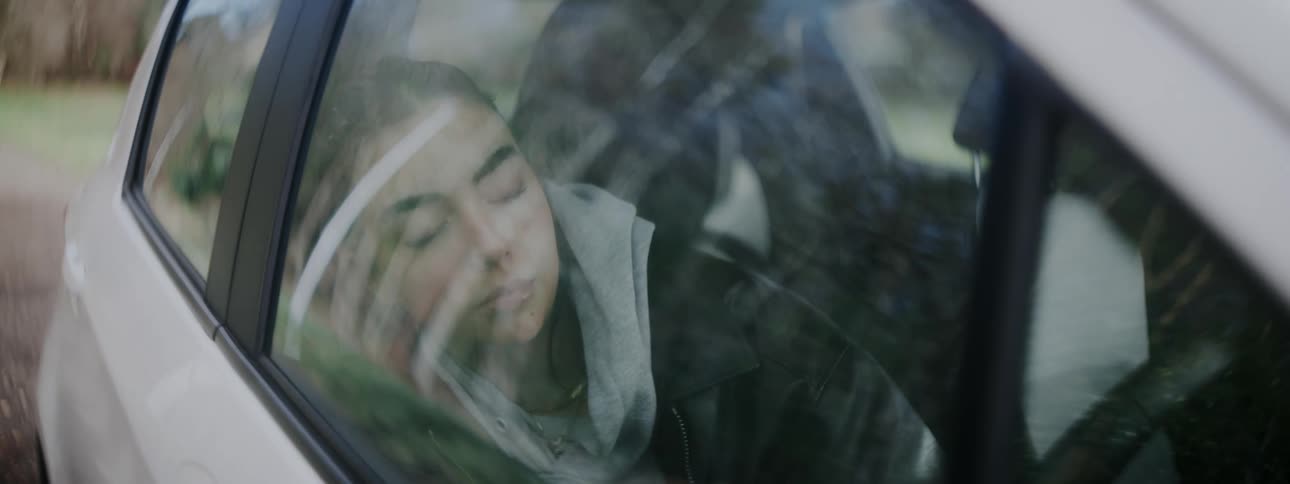 A young woman in a hoodie and jacket is seen through a car window resting her head against the glass with her eyes closed sleeping