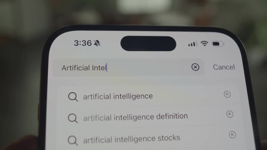 A closeup shot shows a smartphone screen displaying a search bar with the words artificial intelligence and related search suggestions